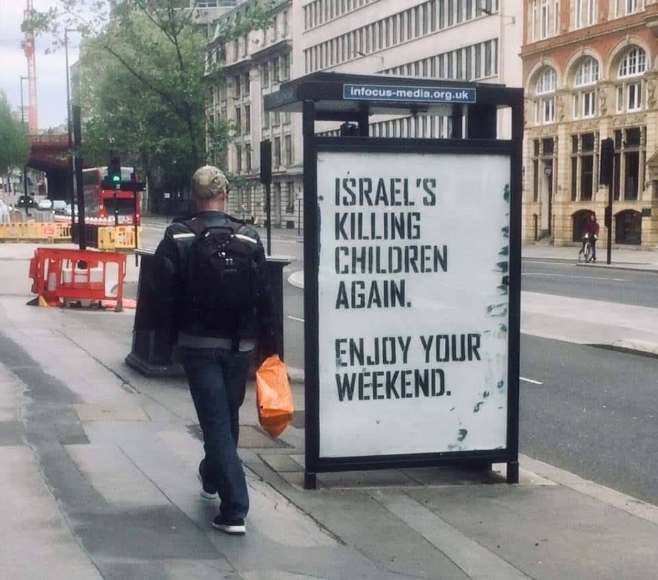 Photo of a street in the UK A person walks past a bus stop with a large sign reading: Israel's killing children again. Enjoy your weekend.